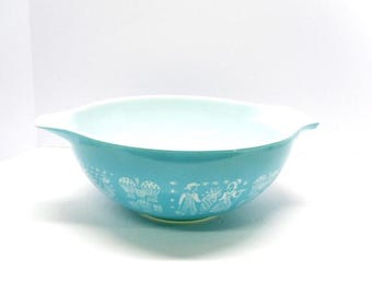 May include: A turquoise Pyrex mixing bowl with a white interior and handles. The exterior features a white, whimsical pattern of figures and objects. The bowl is set against a white background.