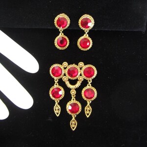 May include: A gold-toned jewelry set featuring a brooch and matching earrings. The brooch has a central design with three large, round, red gemstones, and three dangling gemstones. The earrings each have two red gemstones. The jewelry is set against a black background.