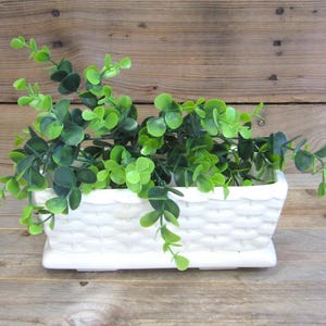 May include: A rectangular white ceramic planter with a molded basket-weave texture on the exterior. The planter is filled with a lush, artificial green eucalyptus plant featuring small, rounded leaves on thin, flexible stems.