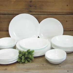 May include: A white dinnerware set, including plates and bowls, arranged on a wooden surface. The set features various sizes of round plates and bowls, perfect for serving meals. A small green plant adds a touch of color.