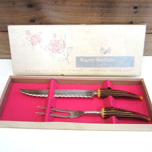 May include: A Regent Sheffield cutlery set in a cream-colored box with rose illustrations. The set includes a serrated knife and a two-pronged fork, both featuring brown, textured, antler-style handles, resting on a bright pink velvet insert.