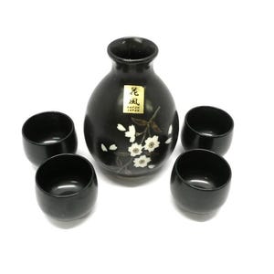 May include: A black ceramic sake set consisting of one rounded bottle and four matching cups. The bottle features a white cherry blossom branch design and a gold rectangular sticker with the text "KAFU JAPAN" and Japanese characters.
