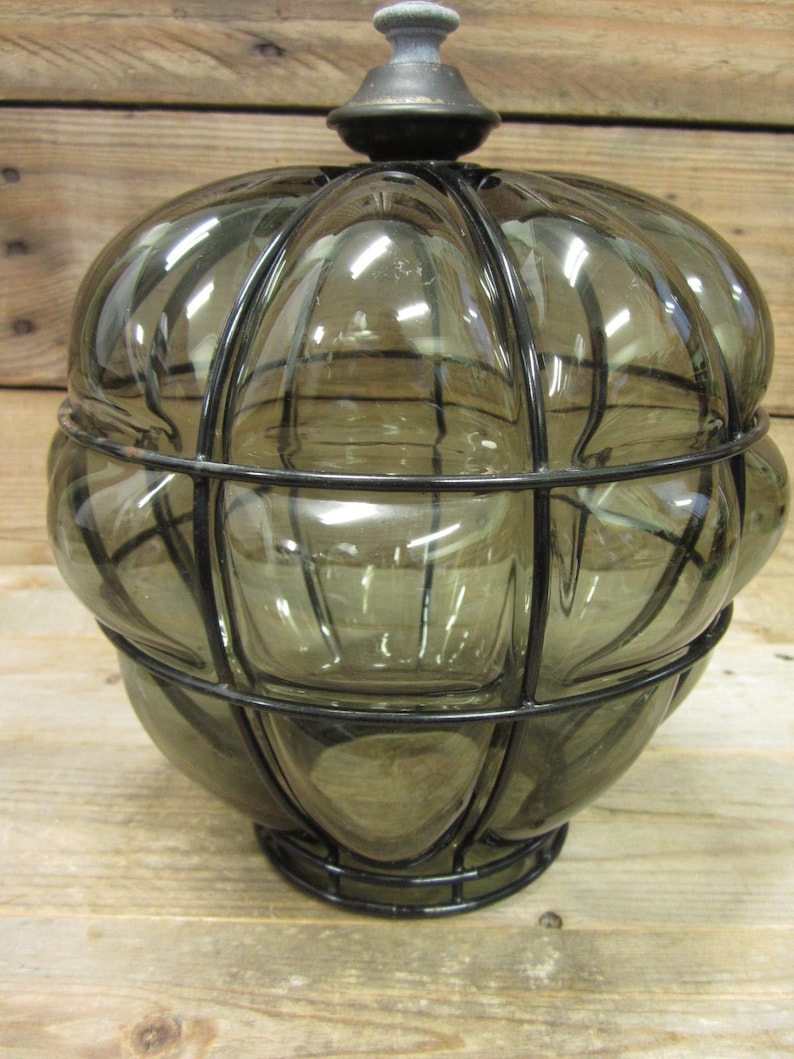 Puede incluir: Un tarro de vidrio decorativo con un tinte gris oscuro, encerrado en una jaula de metal negro. El tarro tiene un dise&ntilde;o redondeado y acanalado y un peque&ntilde;o remate gris oscuro en la parte superior. La base tambi&eacute;n es de metal negro.