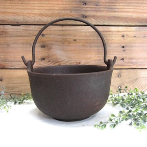 May include: A dark brown cast iron pot with a rounded base and a curved metal handle attached to two side lugs. The exterior surface of the pot has a textured, matte finish, and the handle is positioned in an upright, arched orientation.