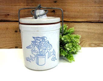 May include: A vintage white ceramic crock with a lid and metal handle. The crock features a blue illustration of a potted plant. The lid has a red rim. The background is a wooden plank wall.