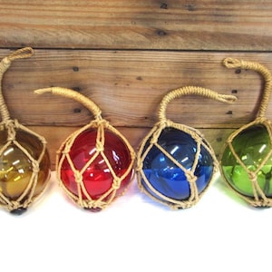 May include: Four spherical glass floats in amber, red, blue, and green, each encased in a decorative tan rope netting. Each glass orb features a sturdy rope handle attached to the top, allowing for hanging display of these colorful nautical items.