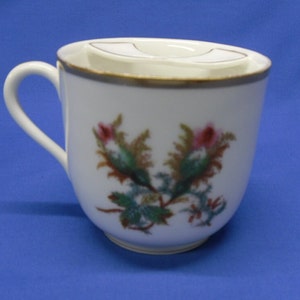 May include: White porcelain teacup with a floral design and a gold rim. The cup has a lid with a small opening for pouring.