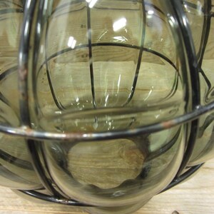 Puede incluir: Un objeto decorativo de vidrio transparente con una jaula de metal oscuro. El vidrio tiene un ligero tinte verde. La jaula de metal tiene un dise&ntilde;o de rejilla. El objeto es probablemente una linterna o un jarr&oacute;n decorativo.