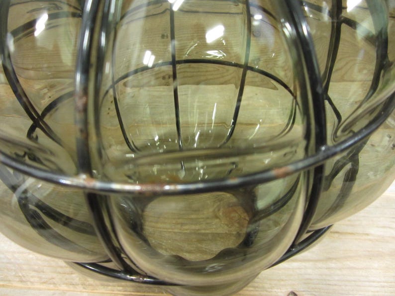 Puede incluir: Un objeto decorativo de vidrio transparente con una jaula de metal oscuro. El vidrio tiene un ligero tinte verde. La jaula de metal tiene un dise&ntilde;o de rejilla. El objeto es probablemente una linterna o un jarr&oacute;n decorativo.