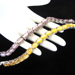 May include: Two link bracelets featuring oval-shaped cabochon stones set in silver-toned metal. One bracelet displays light purple stones with a chatoyant effect, while the other features vibrant yellow stones with a similar cat's eye luster.