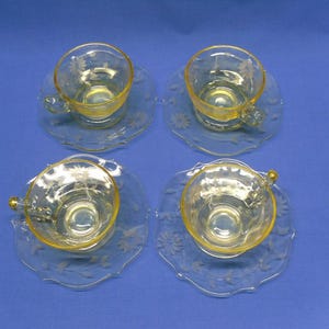 May include: Four vintage yellow-tinted glass teacups and saucers. Each cup sits on a matching saucer with a scalloped edge and floral design. The cups have a delicate handle and a yellow interior. The set is arranged on a blue surface.