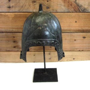 May include: A dark bronze-colored helmet with a pointed top, displayed on a black metal stand. The helmet has a rounded shape with decorative details along the bottom edge. The background is a wooden wall.