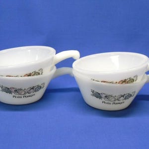 May include: Two white, handled soup bowls stacked on a blue background. Each bowl has a decorative band of vegetables and the words "Petite Potage" printed on the side. The bowls are angled towards the viewer.