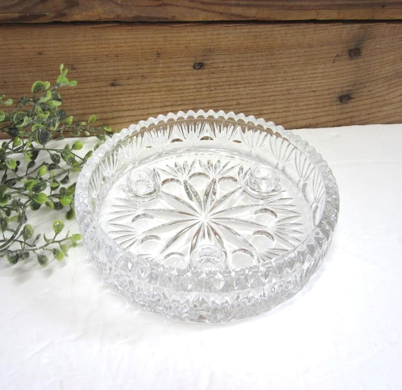 Op de afbeelding: Een heldere, ronde glazen kandelaar met een stervormig ontwerp in het midden. De houder heeft drie kleine kaarsgaten en een decoratieve, getextureerde rand. Het stuk is gemaakt van helder glas en staat op een witte ondergrond.