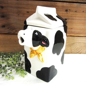 May include: A ceramic cookie jar shaped like a milk carton, painted white with black cow spots. The jar features a cow's face on one side, complete with a pink tongue and a yellow bow tie. The lid is white with black spots.