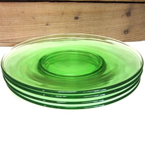 May include: A stack of three circular, translucent green glass saucers. Each saucer features a smooth, flat rim and a slightly recessed center circle. The glass is clear and uniform in color, showing a consistent vibrant green hue throughout.