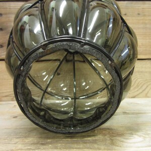 Puede incluir: Una linterna decorativa de vidrio transparente con una jaula de metal negro y una base redonda. La linterna tiene una forma bulbosa con una serie de paneles redondeados. La base tiene un anillo de espuma negra. La linterna est&aacute; sobre una superficie de madera.