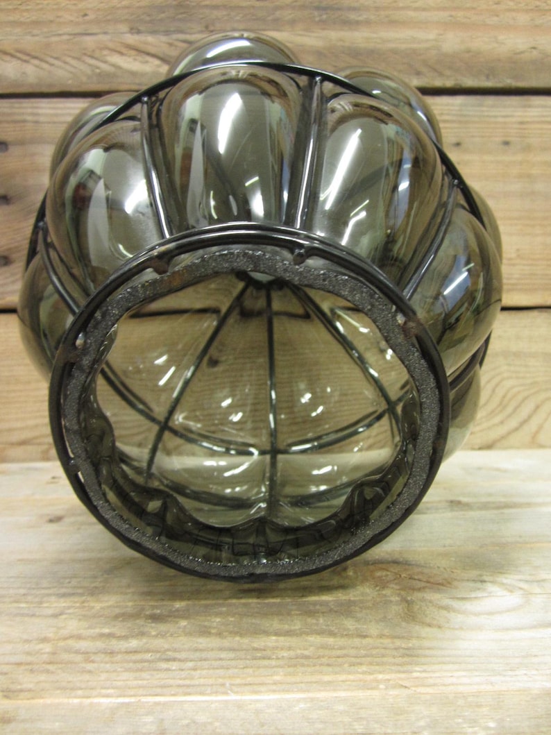 Puede incluir: Una linterna decorativa de vidrio transparente con una jaula de metal negro y una base redonda. La linterna tiene una forma bulbosa con una serie de paneles redondeados. La base tiene un anillo de espuma negra. La linterna est&aacute; sobre una superficie de madera.