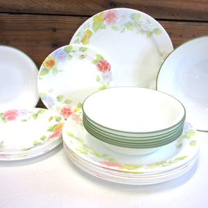 May include: A collection of white dinnerware featuring a delicate floral pattern with pink, yellow, and blue flowers and green leaves. The set includes stacked dinner plates, salad plates, and bowls, some with solid green rims and others with floral borders.