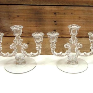 May include: Two clear glass candelabras, each with three candle holders. The candelabras have a decorative, curved design and sit on round bases. The background is a wooden surface.