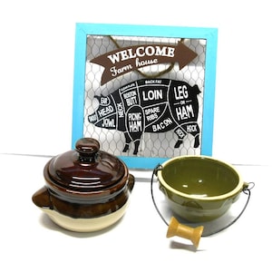May include: A blue and white sign with a pig cut out and labeled with different cuts of meat. The sign says "Welcome Farm house". There are two ceramic bowls, one brown and one green, in the foreground.