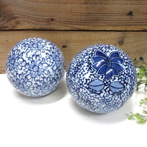 May include: Two decorative ceramic spheres. One sphere features a blue and white floral pattern, while the other has a blue flower design. The spheres are set against a wooden background and a white surface.