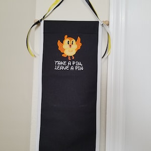 May include: A black fabric pin board with white trim and yellow and black ribbon hanging from the top. The board has a cartoon bird with the text "Take a pin, leave a pin" embroidered on it.