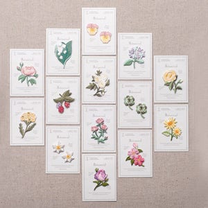 Op de afbeelding: Een set van 15 strijkbare patches met verschillende botanische ontwerpen. De patches zijn wit met roze, gele en groene borduursels. Elke patch heeft de tekst "Botanical" erop gedrukt.