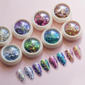 May include: Eight small round white containers filled with multi-colored iridescent nail glitter flakes in various shades including silver, blue, purple, green, and gold, accompanied by eight clear nail tips displaying the glitter applied to them.