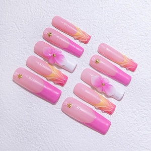 Könnte beinhalten: Ein Set aus zehn künstlichen Nägeln in verschiedenen Designs. Die Nägel weisen einen Farbverlauf von Rosa und Weiß auf, einige sind mit 3D-Blumen und goldenen Sternakzenten verziert. Die Nägel sind auf einer weißen Oberfläche angeordnet.