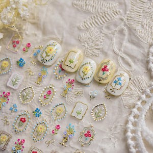 May include: A collection of nail art stickers featuring various floral designs, including yellow daisies, red roses, and blue forget-me-nots, framed in gold metallic borders. The set includes individual flower motifs and small bows on a clear backing.