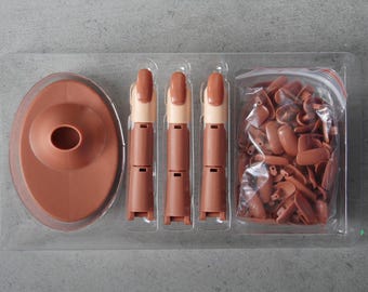 May include: A nail art kit in a clear plastic tray. The kit includes a brown oval base, three articulated practice fingers with tan and brown nails, and a bag of brown nail tips. The items are arranged on a gray surface.