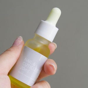 Pode incluir: Uma pequena garrafa de vidro transparente de óleo para unhas e cutículas, segurada por uma mão. A garrafa tem um rótulo branco com o texto "NAIL & CUTICLE OIL 20ml". O óleo dentro é amarelo. A garrafa tem um conta-gotas branco.