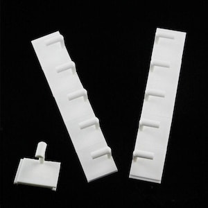 May include: White plastic shelf supports and shelf clips against a black background. The shelf supports are long, rectangular strips with evenly spaced, cylindrical pegs. A smaller, square clip with a single peg is also present.