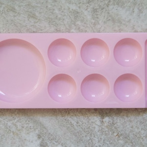 Pode incluir: Paleta de pintura de plástico rosa claro com um grande poço circular, seis poços circulares menores e um poço retangular. A paleta é projetada para misturar tintas e é ideal para artistas e artesãos.