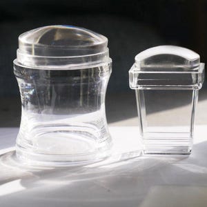 May include: Two clear, transparent glass objects. One is a jar-shaped container with a curved body and a domed lid. The other is a rectangular, box-like container with a curved top. Both are on a white surface.