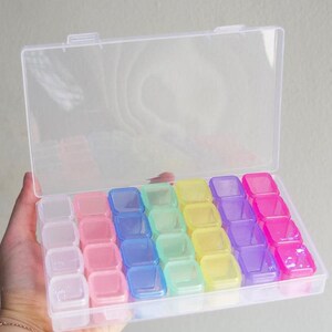 May include: A clear plastic storage box with a hinged lid, filled with small, colorful compartments. The compartments are arranged in rows and are various colors, including pink, blue, green, yellow, purple, and clear.