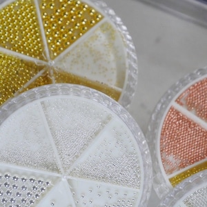 Puede incluir: Un conjunto de recipientes redondos de plástico transparente divididos en compartimentos triangulares, llenos de diminutas cuentas metálicas en acabados dorado, plateado, oro rosa y blanco, para manualidades o arte de uñas.