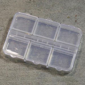 May include: A clear plastic organizer with six individual compartments, each with a hinged lid. The compartments are numbered 1 through 6. The organizer is designed to hold small items and is shown on a gray surface.