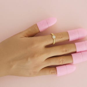 Può includere: Una mano con un anello d'oro con una pietra bianca centrale e protezioni per le dita rosa. L'anello è sull'anulare. Le protezioni per le dita rosa sono sulle altre dita. Lo sfondo è rosa pieno.