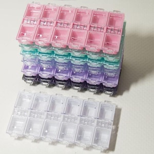 May include: A collection of clear plastic pill organisers in various colours. The stack includes pink, teal, purple, and black organisers. A separate, clear organiser is in front. Each organiser has individual compartments with hinged lids.