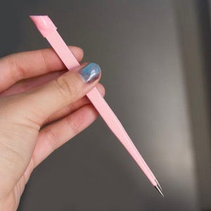 Può includere: Uno strumento per unghie rosa chiaro con una punta e un'estremità piatta e angolata. Lo strumento è tenuto in mano su uno sfondo neutro. Questo strumento per manicure viene utilizzato per la cura delle unghie.