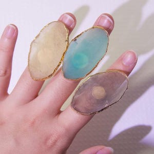Op de afbeelding: Drie edelsteenringen met goudkleurige randen worden getoond. De ringen hebben ovaalvormige stenen in tinten geel, blauwgroen en grijs. De ringen zitten op een hand met een lichtpaarse achtergrond.