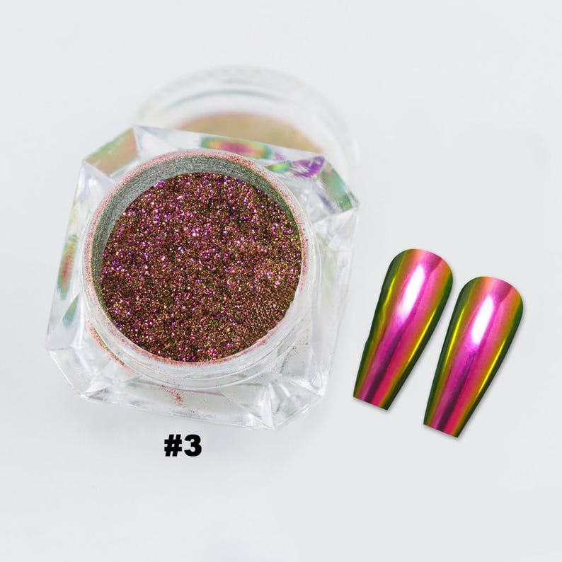May include: A clear jar containing shimmering, reddish-brown nail powder, marked with the number "#3". Two iridescent, oval-shaped nail tips are to the right. The tips reflect a gradient of pink, yellow, and green.