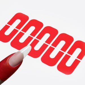 May include: A red nail with a white tip is positioned near a red nail art stencil on a white surface. The stencil design features a series of connected, open rectangles.