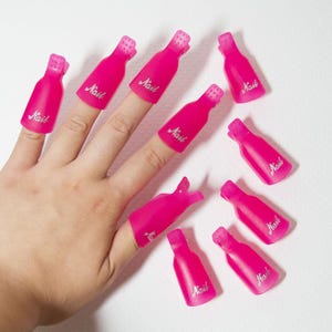Può includere: Clip rosa acceso per la rimozione dello smalto con la scritta "Nail" stampata sopra. Le clip sono progettate per essere posizionate sulle dita per una facile rimozione dello smalto. L'immagine mostra diverse clip, alcune sulle dita e altre sparse.