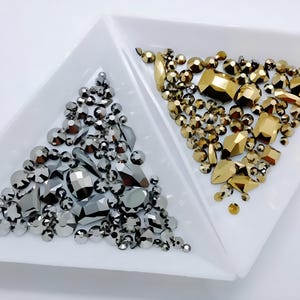 May include: A collection of faceted rhinestones in two white triangular trays. The left tray contains metallic silver and dark gray stones in various shapes, while the right tray holds metallic gold and bronze stones in assorted geometric cuts.