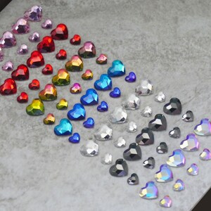 May include: A collection of faceted heart-shaped rhinestones in a gradient of colors including pink, red, yellow, blue, silver, black, and iridescent finishes. The small, decorative gems are arranged in rows, showcasing various sizes and reflective surfaces.