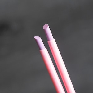 Peut inclure: Deux crayons cosmétiques roses avec des pointes lavande sont inclinés sur un fond gris foncé. Les crayons sont cylindriques et semblent neufs. Les pointes sont d'une nuance de lavande plus claire que le corps du crayon.