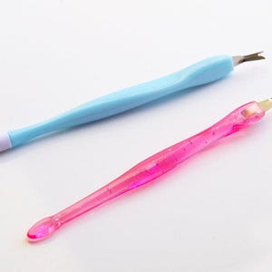 May include: Two nail cuticle tools. One is light blue with a white tip and a metal blade. The other is pink with glitter and a metal blade. Both tools are on a white background.
